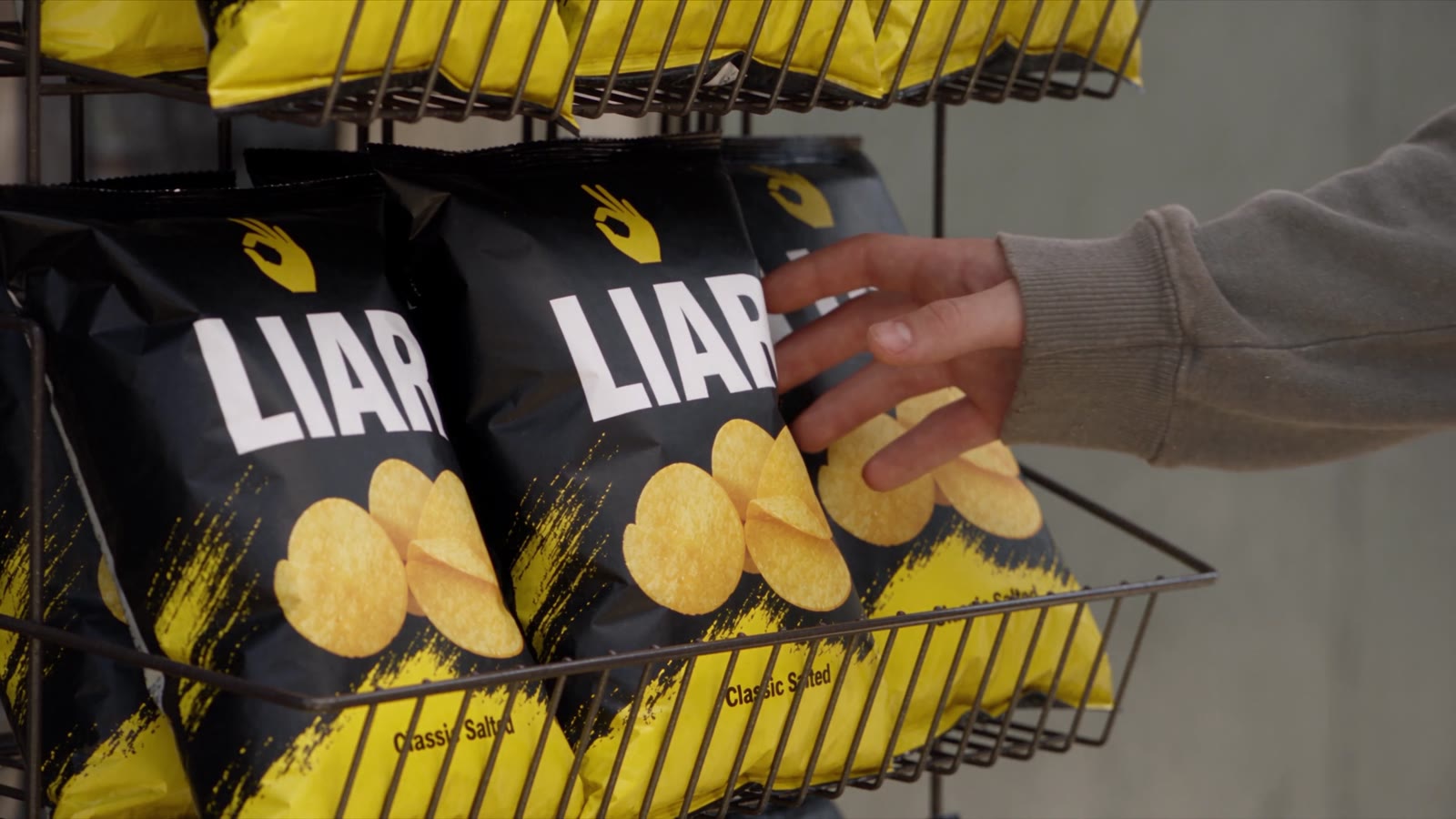 A hand reaching for a bag of LIAR chips on a kiosk rack